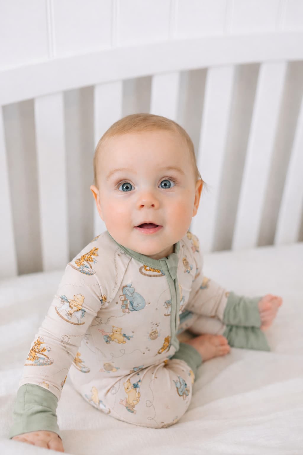 Baby in bamboo sleeper pajamas with Pooh Bear prints sitting in a crib.
