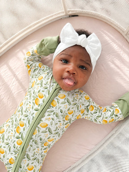 A baby wearing a bamboo sleeper with a yellow and green lemon print, a white bow headband, and light green mittens.