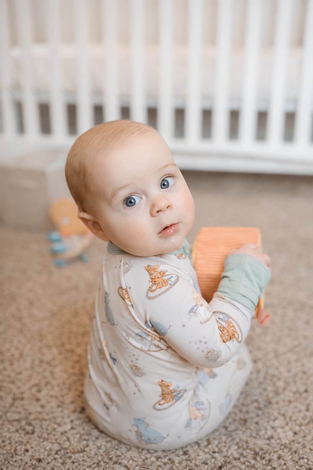 Baby wearing a classic pooh bamboo sleeper sitting on a carpeted floor holding a toy, with a crib in the background.