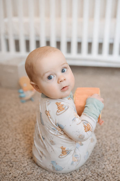 Baby wearing a classic pooh bamboo sleeper sitting on a carpeted floor holding a toy, with a crib in the background.