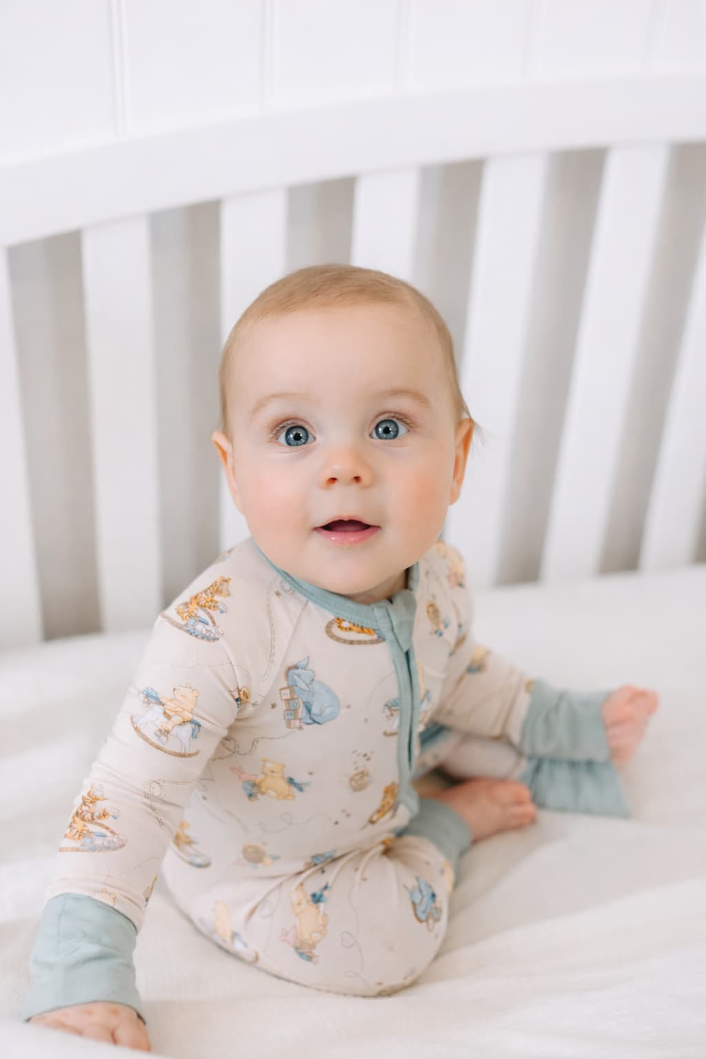 Baby in a crib wearing a Classic Pooh bamboo sleeper pajamas featuring Pooh Bear, Eeyore, Tigger, and Piglet with vinage toys. 