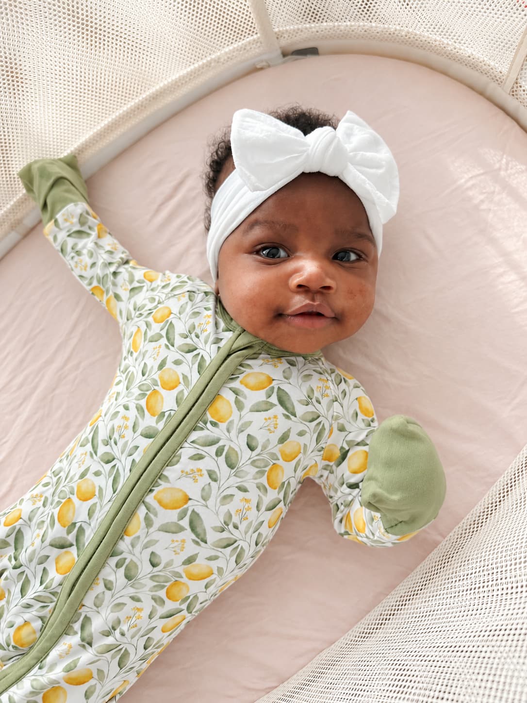 A baby lying on a pink bassinet wearing a white bow headband and a light green and white lemon-patterned bamboo sleeper.