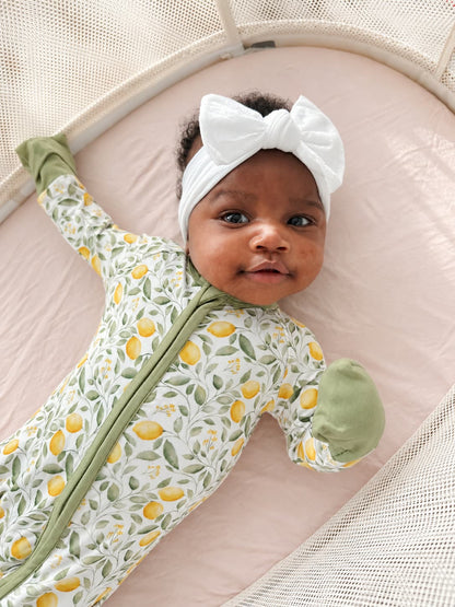 A baby lying on a pink bassinet wearing a white bow headband and a light green and white lemon-patterned bamboo sleeper.