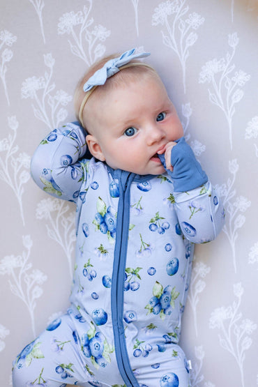 A baby lying on a floral-patterned blanket, wearing a light blue zip-up sleeper with a blueberry print and a matching blue bow headband.