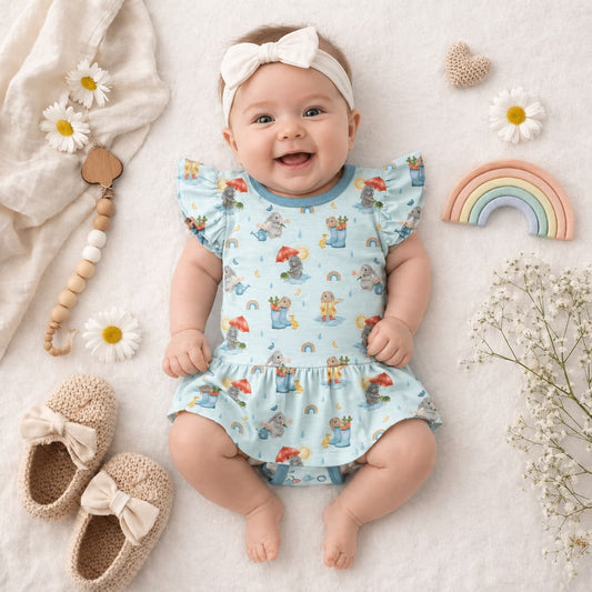 Baby in a blue bamboo romper with bunny and rainbow print surrounded by toys and flowers on a light background.