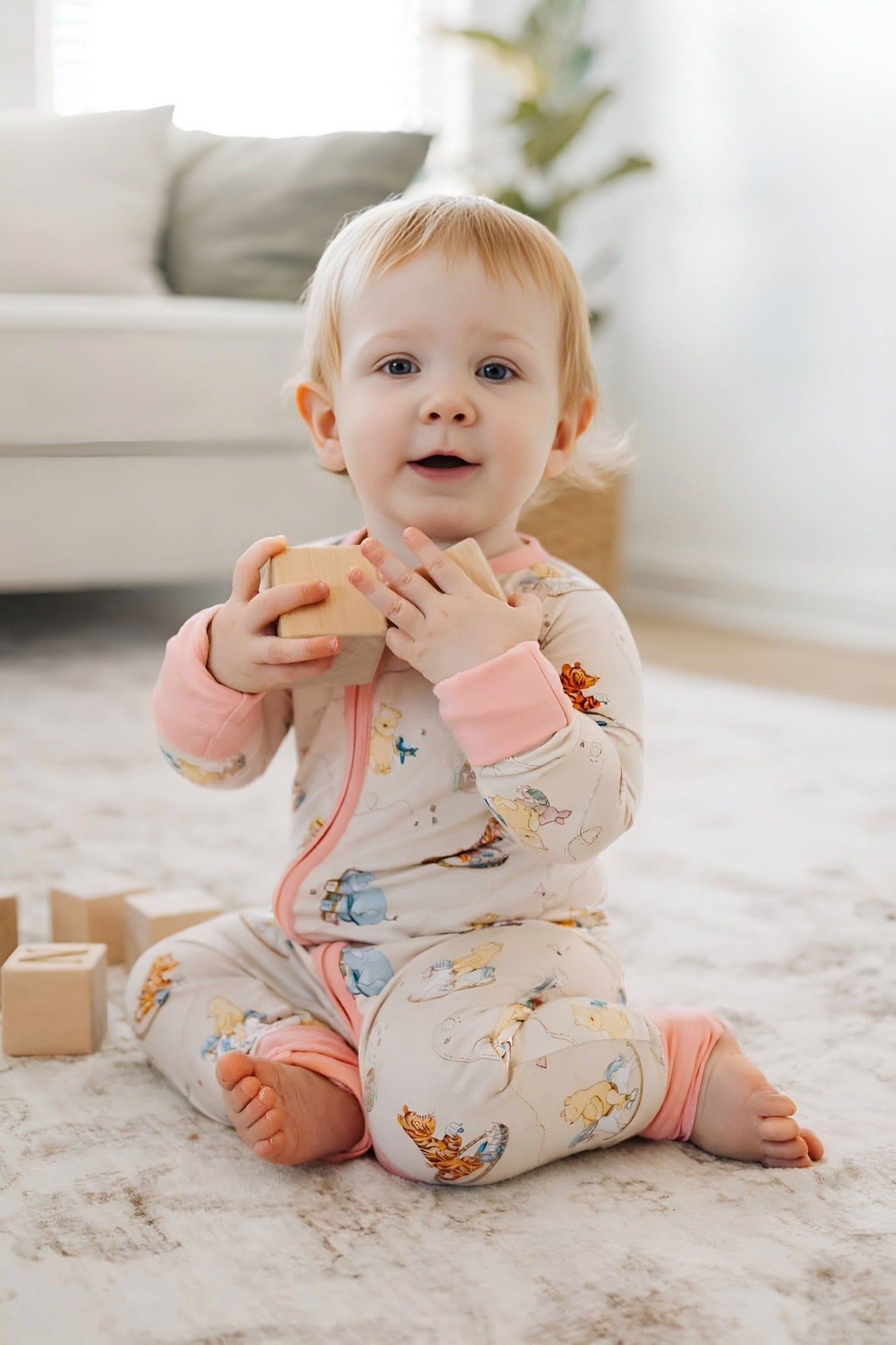 Baby in classic Pooh bamboo pajams sitting on a carpeted floor holding wooden blocks in a living room.