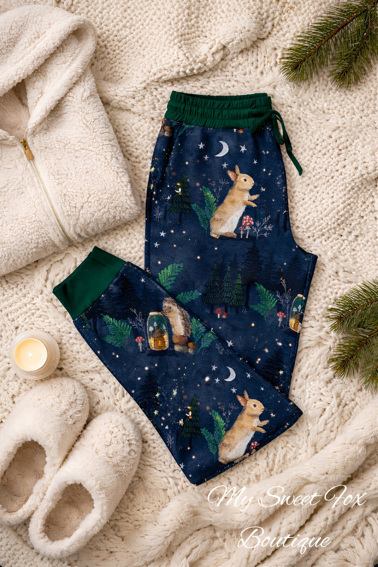 Navy blue woodland print pajama pants with bunny, hedgehog, firefly design on a soft surface with slippers and a blanket.