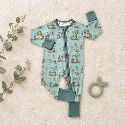 Baby bamboo sleeper pajamas with animal pattern on a beige surface with a green teething ring and leaves.