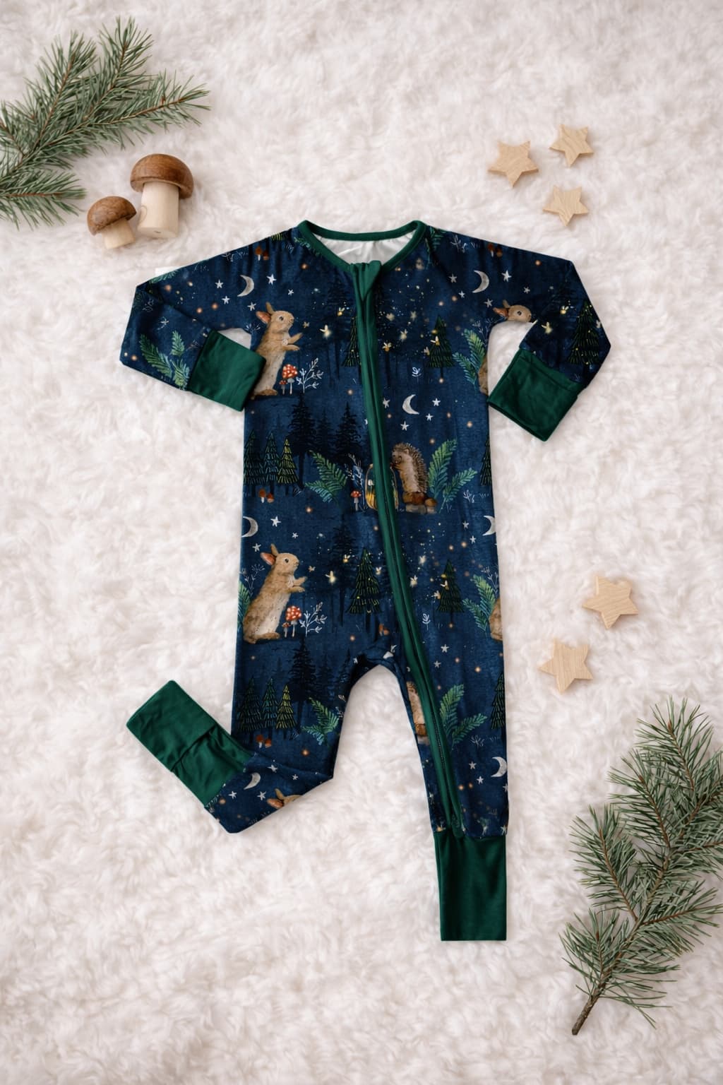 Baby bamboo pajamas with hedgehogs, bunnies, mason jars, fireflies, stars, acorn house, and woodland theme pattern on a blanket with wooden decors and pines.