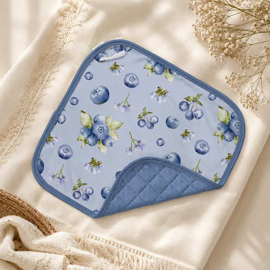 A bamboo baby lovey or baby washcloth with a blueberry print, folded to show its quilted blue underside, styled on a cream-colored blanket with dried flowers.