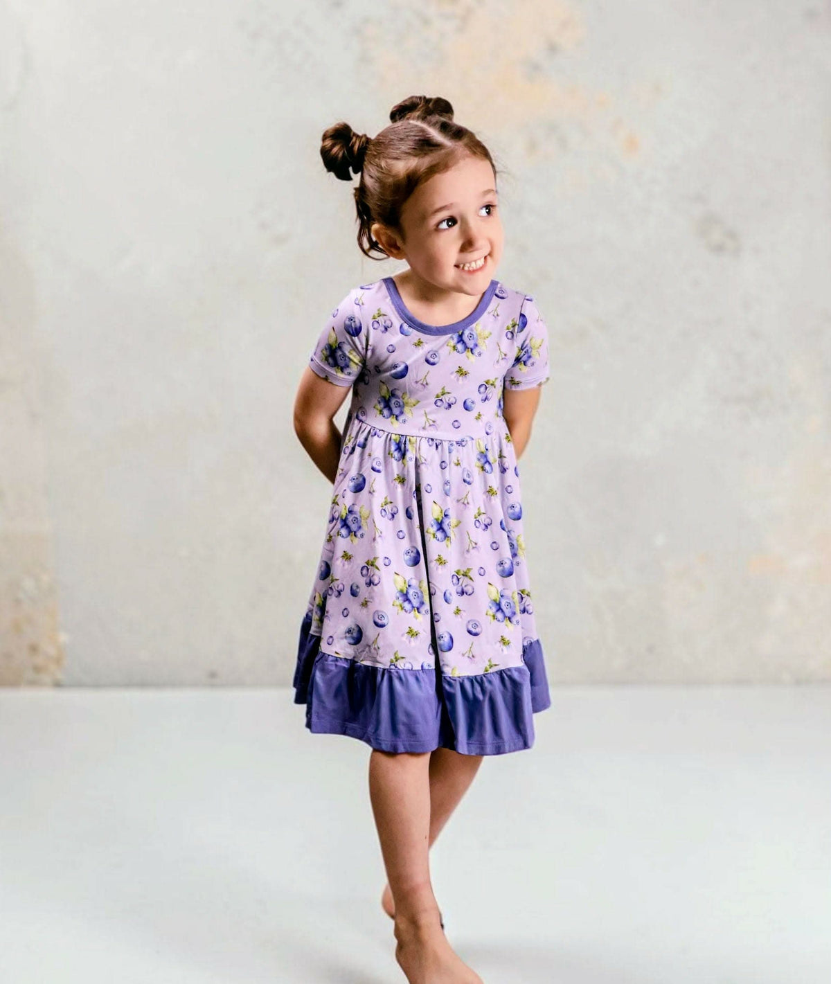 A young girl with her hair in two buns stands barefoot, wearing a blueberry bamboo twirl dress. The dress has flutter sleeves and solid blue ruffled trim at the hem, set against a textured light gray wall.