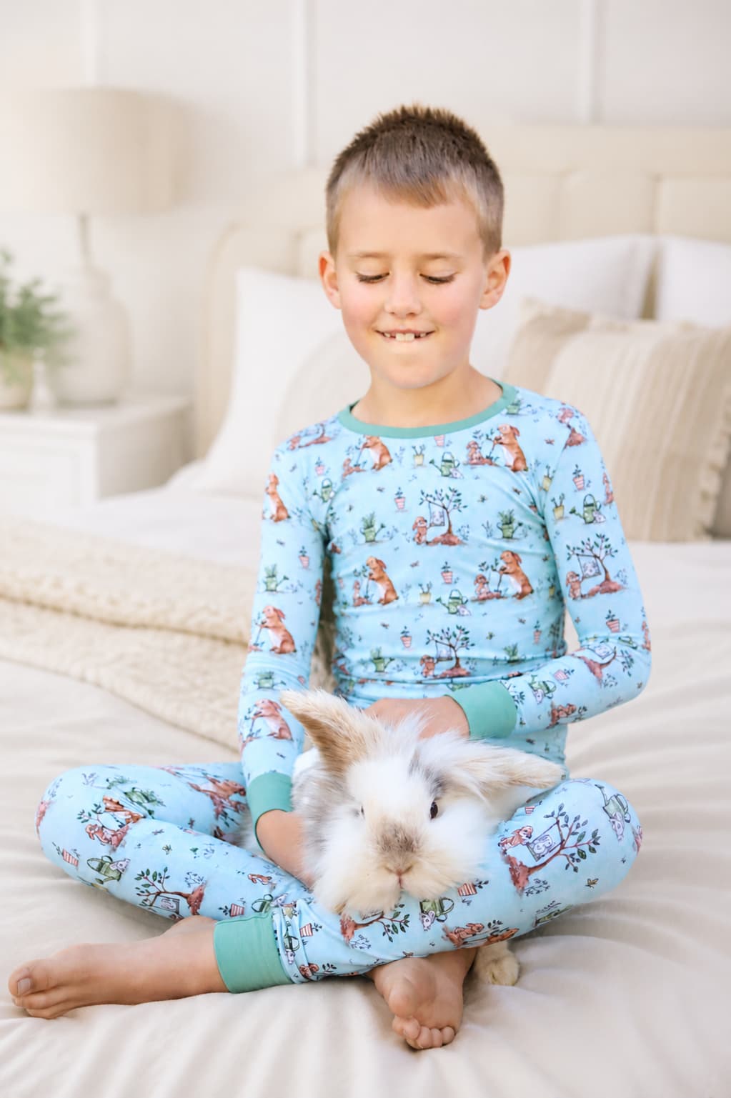 Boy wearing a blue bamboo pajamas set with bunny and garden print, while holding a small white bunny.