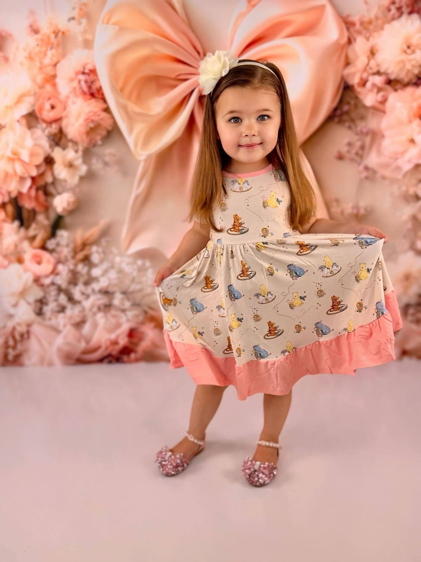Young girl in a Classic Pooh bamboo twirl dress with blush ruffles standing against a floral pink backdrop