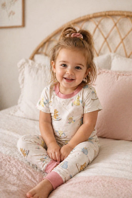 Child wearing Classic Pooh bamboo pajamas sitting on a bed with pink pillows and a wicker headboard.