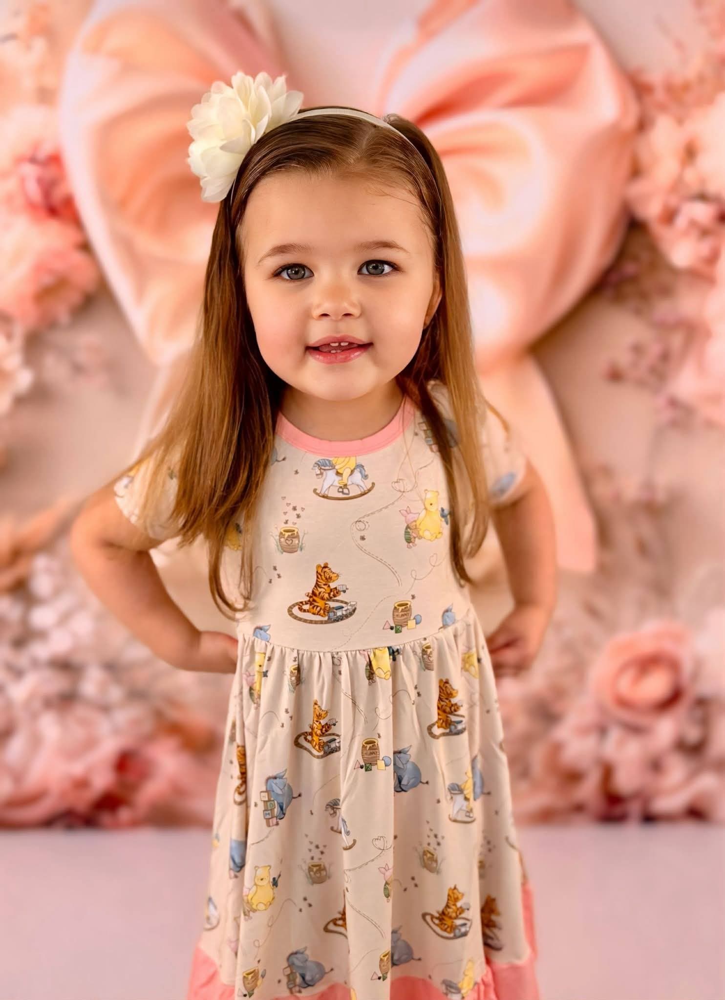 Young girl wearing a Winnie the Pooh bamboo twirl dress with a floral headband, standing against a decorative pink and white floral background.