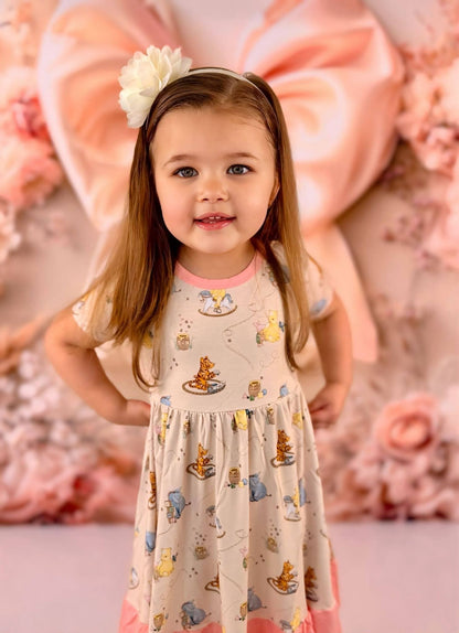 Young girl wearing a Winnie the Pooh bamboo twirl dress with a floral headband, standing against a decorative pink and white floral background.