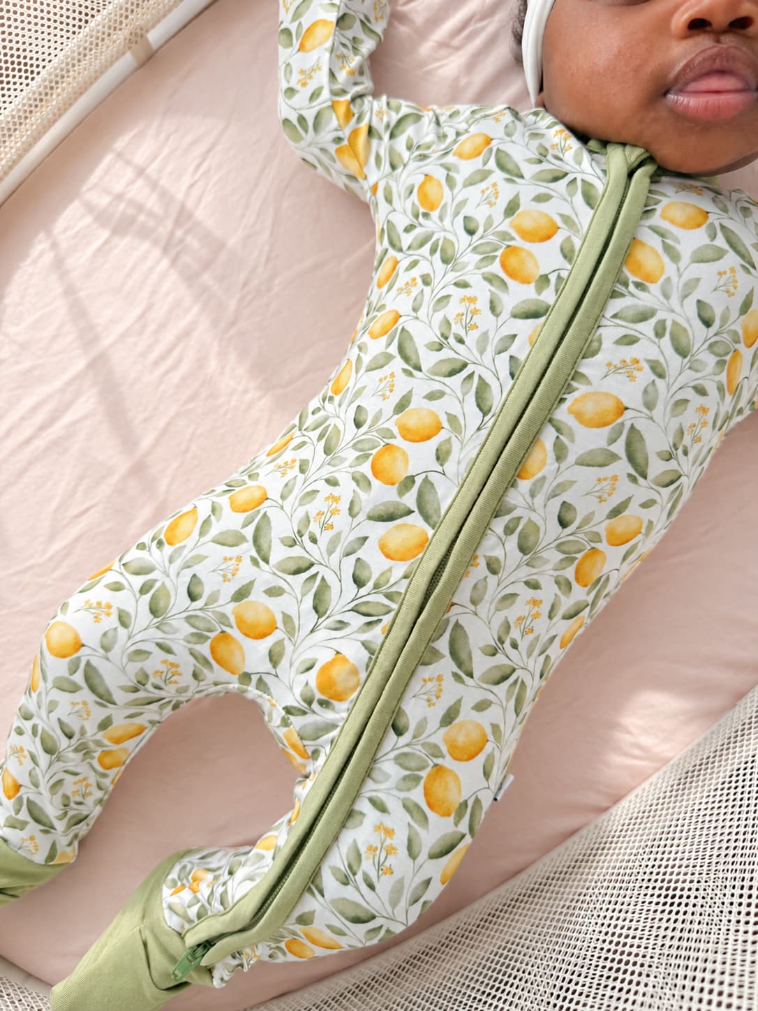 A baby lying on a pink mattress wearing a bamboo footed zip-up sleeper with a white background, green leaf pattern, and yellow lemon print. The bamboo sleeper has sage green trim along the zipper and feet.