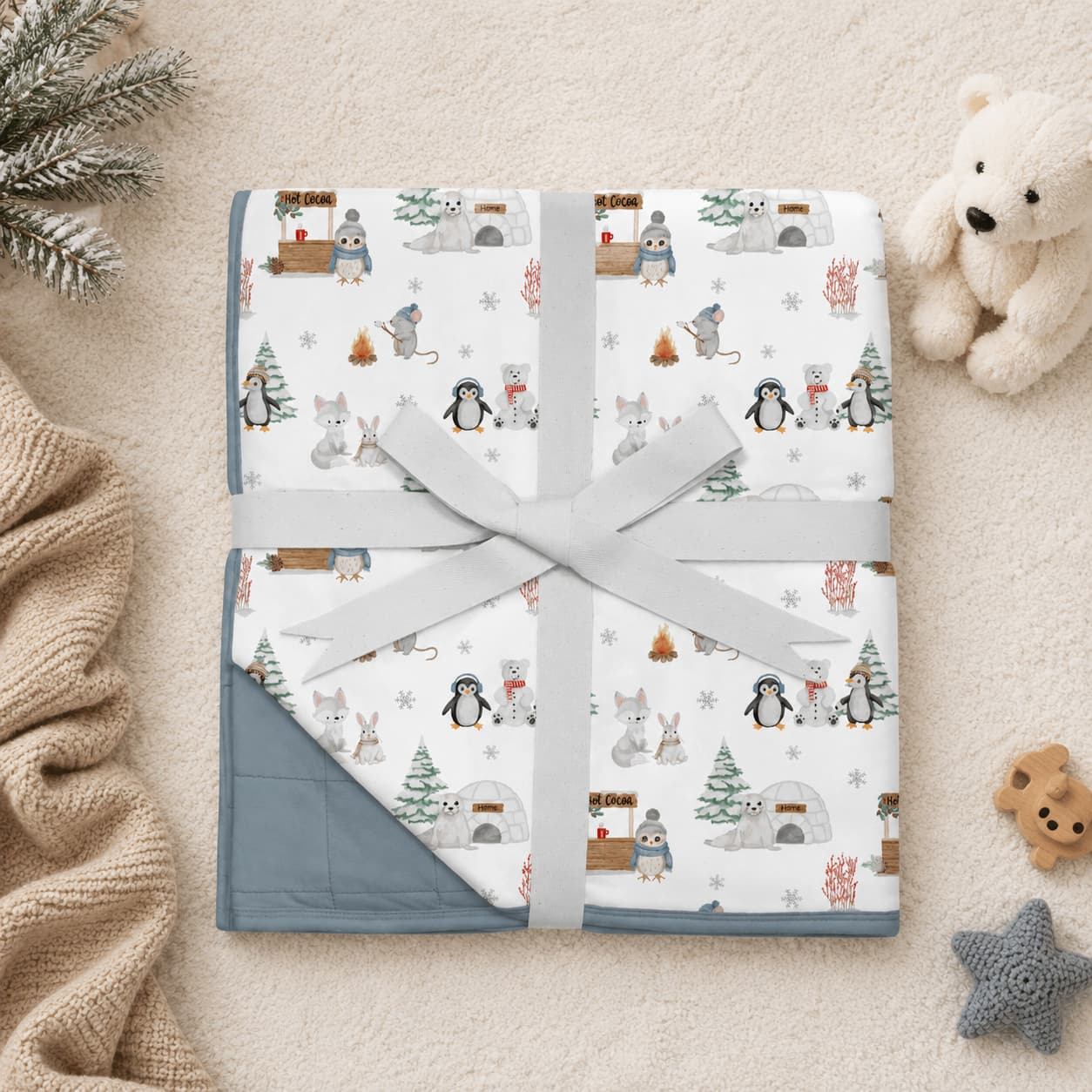 Folded bamboo baby blanket with a winter animal print and a solid blue reverse side, wrapped in a light gray ribbon and tied in a bow, presented on a beige textured blanket with cozy props like a teddy bear and knit sweater.