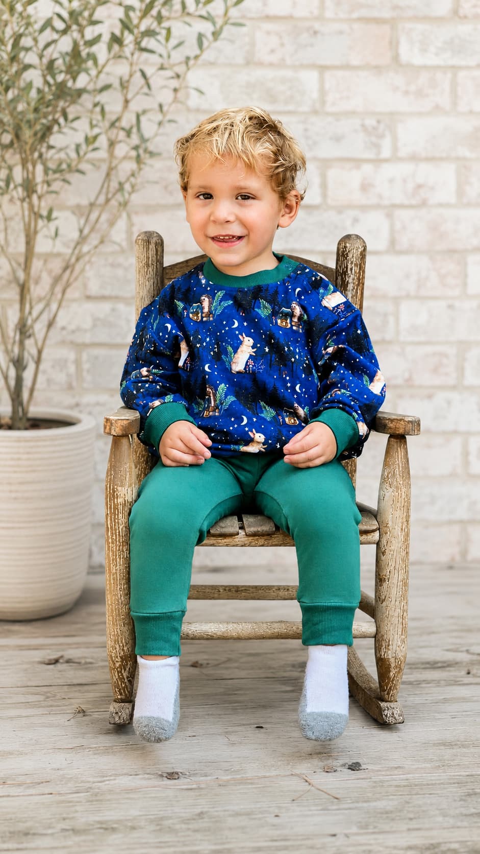 Child sitting on a wooden chair wearing a navy blue woodland printed sweatshirt and green french terry bamboo jogger set. 
