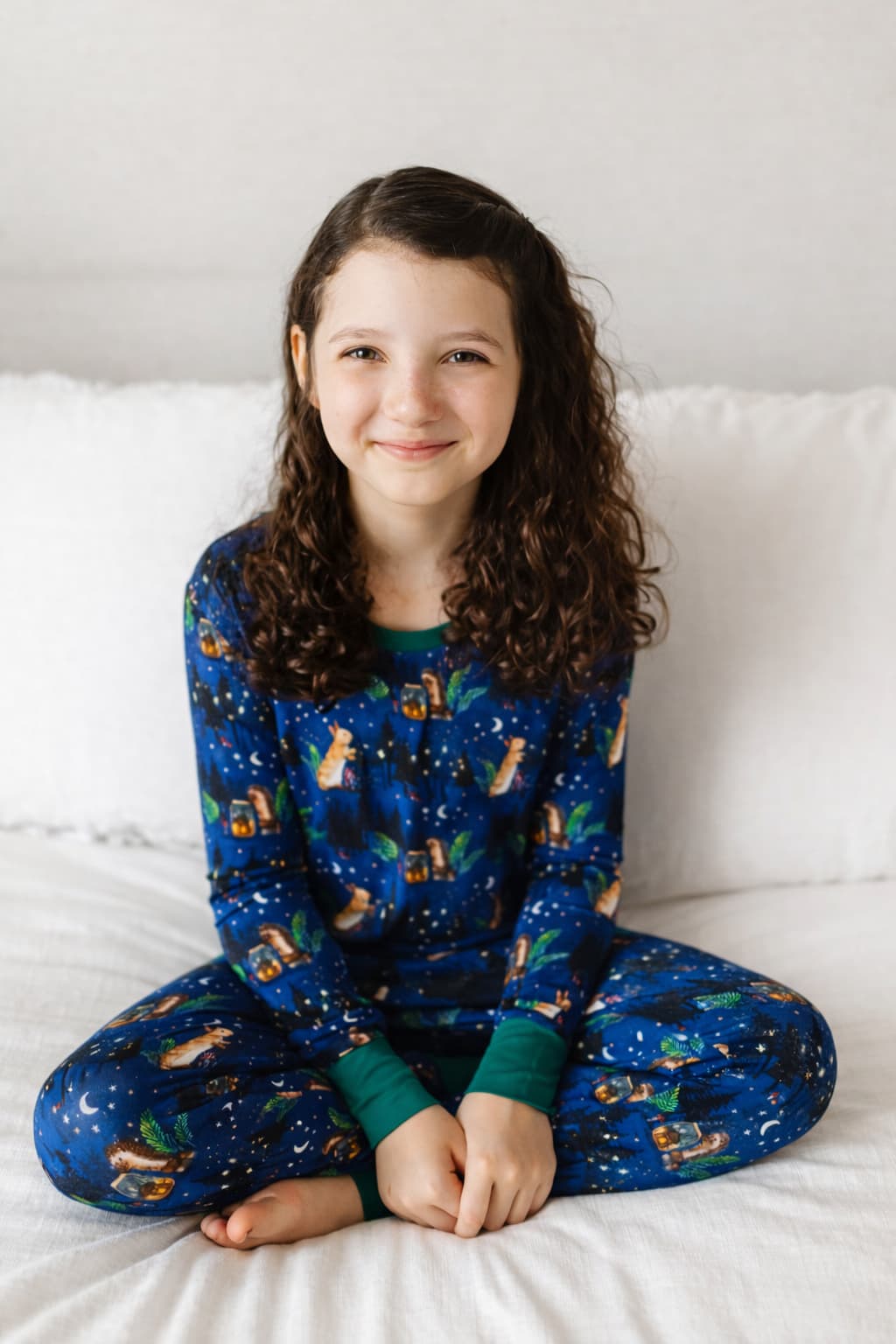 Young girl wearing a blue woodland themed bamboo pajama set sitting on a white bed.