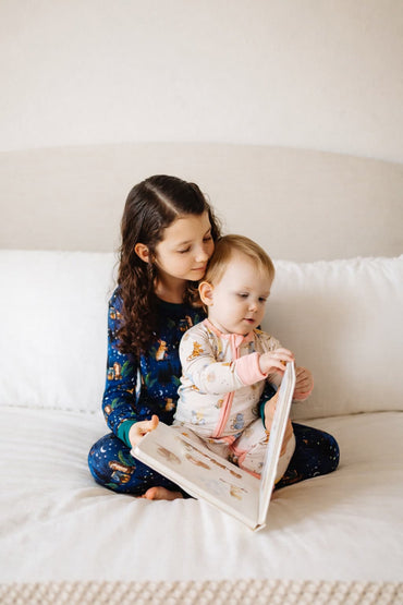 Two children sitting on a bed, reading a book together in their bamboo pajamas.