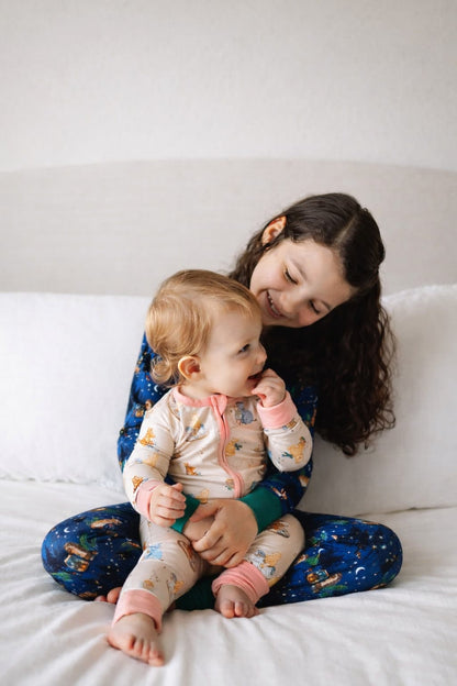 Sister and baby sister sitting on a bed wearing bamboo pajamas. One bamboo pajamas has classic pooh. The other has woodland print.
