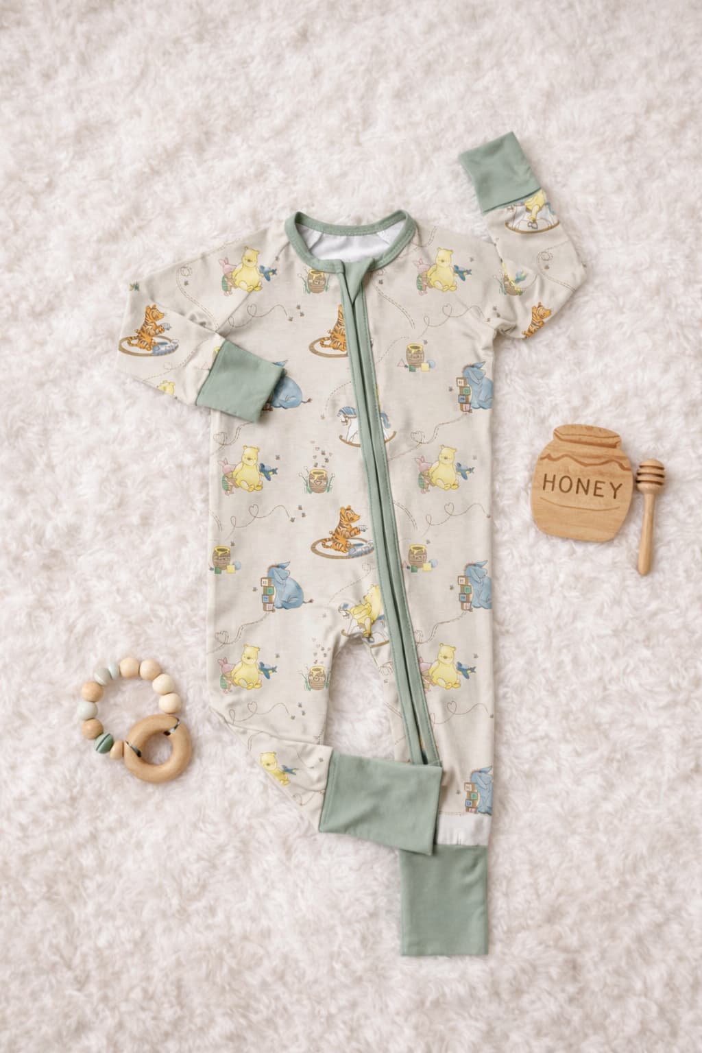 Bamboo baby sleeper pajamas featuring classic pooh and friends placed on a fluffy blanket with wooden teether and a wooden sign that says honey. The sleeper has soft sage zipper and converible hands and feets cuffs.