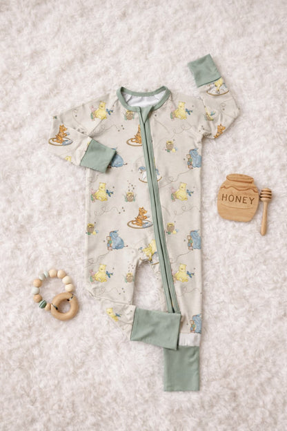 Bamboo baby sleeper pajamas featuring classic pooh and friends placed on a fluffy blanket with wooden teether and a wooden sign that says honey. The sleeper has soft sage zipper and converible hands and feets cuffs.