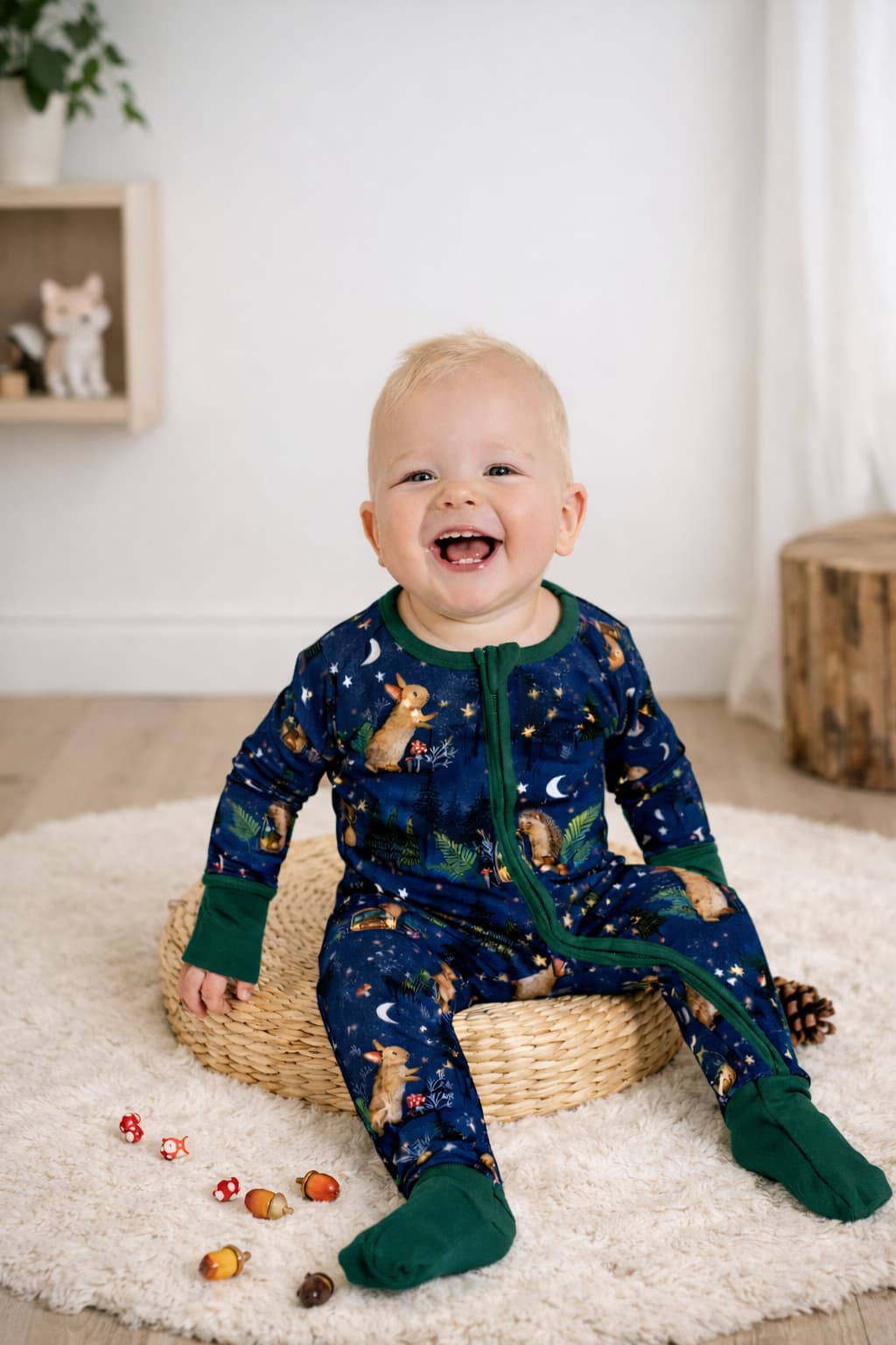 Baby in a navy blue pajama sitting on a white rug. The print has hedgehogs, bunnies, mason jar, acorn houses, fireflies, stars, and a background with trees. 