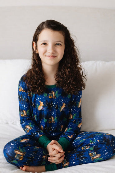 Young girl wearing a blue bamboo pajama set with woodland animal pattern on a white bed.